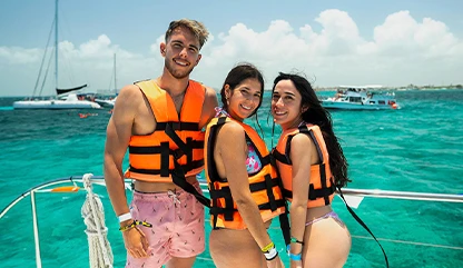 Two women one man in orange lifejackets on a Cancun boat tour with blue ocean blue sky and isla mujeres yachts sailing.