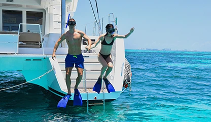 Man in blue shorts woman in black bikini jumping off a luxury Cancun catamaran into the blue ocean to snorkel near Isla Mujeres.