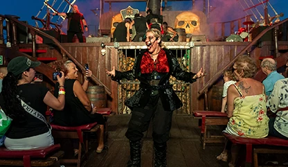 Group of tourists inside the pirate ship sitting in wooden benches recording and listening to a female pirate with a fun appearance.