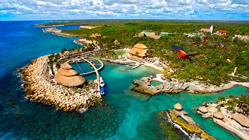 Breathtaking aerial view of the entire Xcaret eco-park, with red macaws soaring over the tropical landscape and crystal-clear waters.