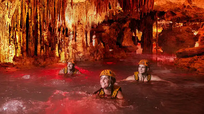 Group of friends swimming in the underground river at Xplor Fuego eco-park, wearing life jackets and helmets with an orange ilumination of the cave.